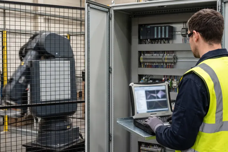 A controls engineer at an open cabinet of PLC modules beside a guarded industrial robot cell, working on a rugged laptop