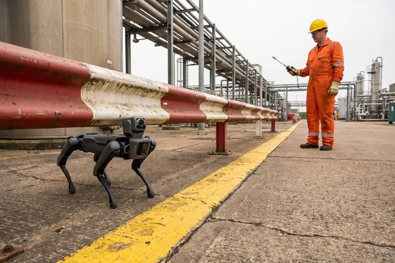A four-legged robot waiting at a painted boundary line at the edge of a process area while a technician checks the air with a gas detector.