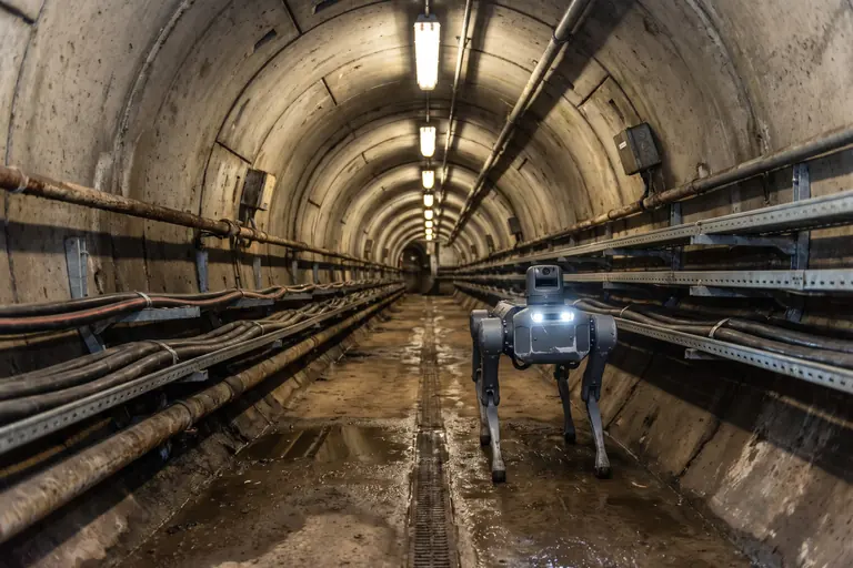 A four-legged inspection robot with its lights on walking down a concrete cable tunnel lined with power cables on steel racks.