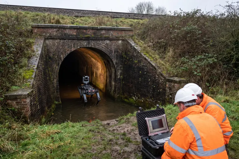 A four-legged inspection robot walking into a brick railway culvert through shallow water while two workers in hi-vis watch a laptop on the bank outside.