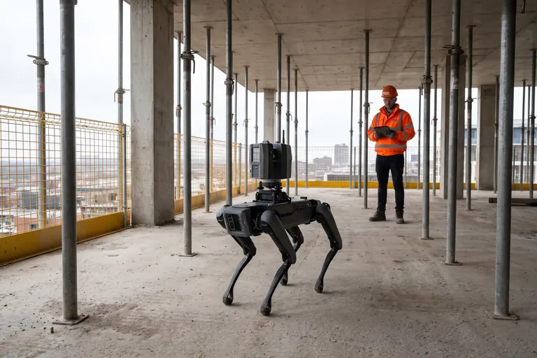 A four-legged robot carrying a laser scanner on an upper floor of a concrete frame under construction, watched by a site engineer.