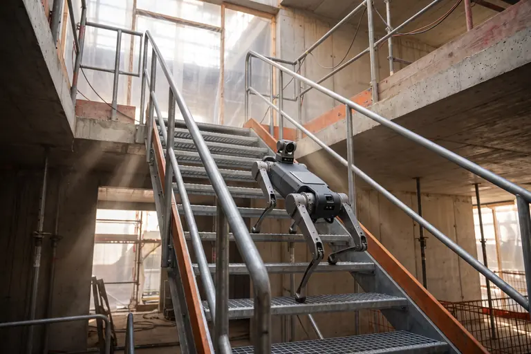 A four-legged robot climbing a temporary steel stair between floors of a building under construction.