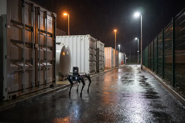 A four-legged inspection robot with its lights on walking past a row of containerised generators in a data centre yard at night.