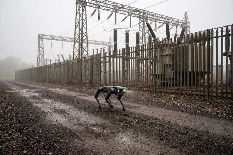A four-legged inspection robot walking a gravel route through a misty outdoor substation compound, past gantries and transformers.