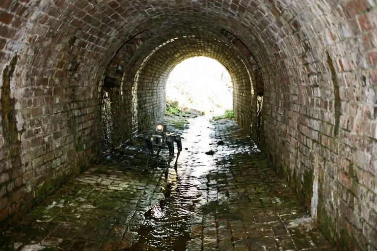 A small legged robot walking through shallow water inside a narrow brick culvert towards its bright entrance.