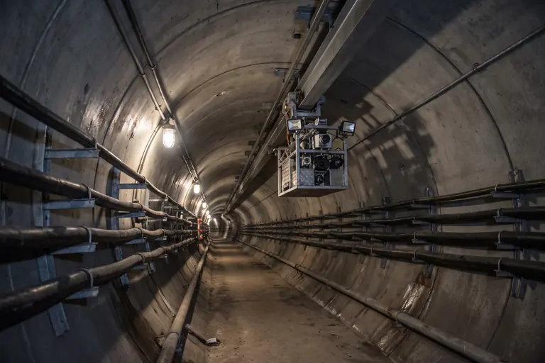A compact inspection vehicle hanging from a monorail at the crown of a long, deep cable tunnel.