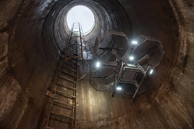 A caged inspection drone with bright lights descending a deep circular concrete shaft, seen from below.