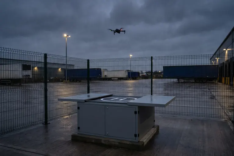 A security drone returning at dusk to an open drone dock inside the fence of a UK logistics yard